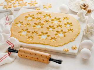 Rolled out dough with cut out Christmas gingerbread snowflake shapes with baking ingredients and with rolling pin on white kitchen table. Top view. Christmas baking concept background.