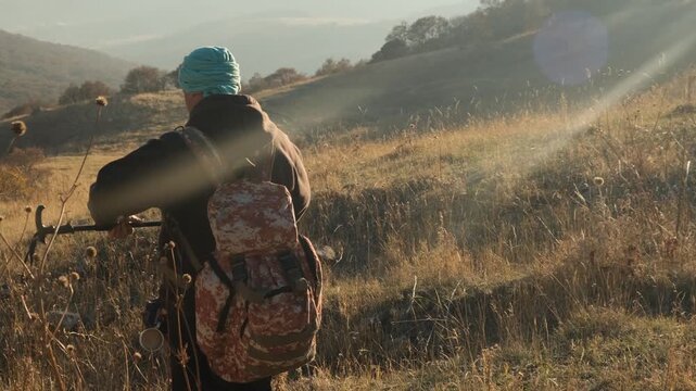 A male traveler with a backpack walks down a mountain slope at sunset holding a telescopic trekking pole and adjusting its length. Using hiking poles in the mountains. Solo hiking adventure.