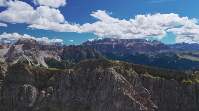 Drone aerial view revealing the sharp ridges of Seceda in the Dolomites, Italy. Wide panoramic landscape with rocky peaks, soft clouds, and clear blue sky on a sunny day