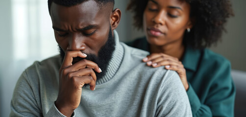 Black man sitting with closed eyes, hand on chin, looking sad. Woman comforts him, hand on shoulder, eyes closed. Man stressed, woman showing empathy and support. Couple in emotional moment together.
