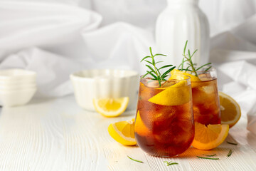 Iced tea or alcoholic cocktail with ice, rosemary and lemon slices on the white table.
