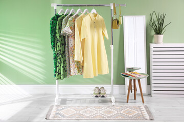 Different women's clothes hanging on rack, mirror, side table and houseplant near green wall indoors