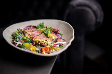 A gourmet salad of seared duck breast, mixed greens, pistachio nuts, and orange slices is served in a modern bowl against a dark background, showcasing fine dining