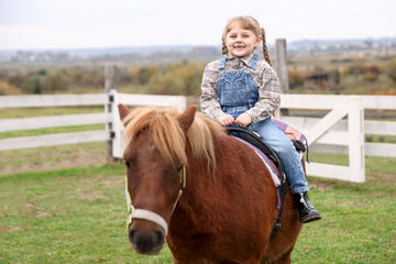 Fototapeta premium Equine assisted therapy. Little girl riding beautiful pony outdoors. Lovely domesticated pet
