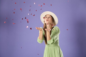 Happy woman blowing heart shaped confetti on violet background