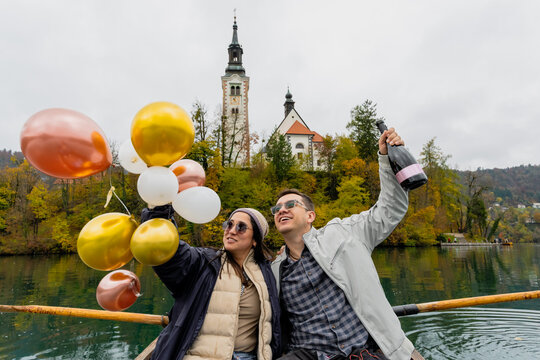 Young couple celebrating their engagement with balloons and champagne outdoors at Lake Bled surrounded by autumn foliage
