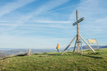 Gustav-Ströhmfeld-Weg: Gipfelkreuz mit Ruhebank am ehemaligen Vulkanschlot  Jusiberg in Kohlberg vor blauer Mauer der schwäbischen Alb mit drei Kaiserbergen im Hintergrund vor blauem Himmel mit Wolken