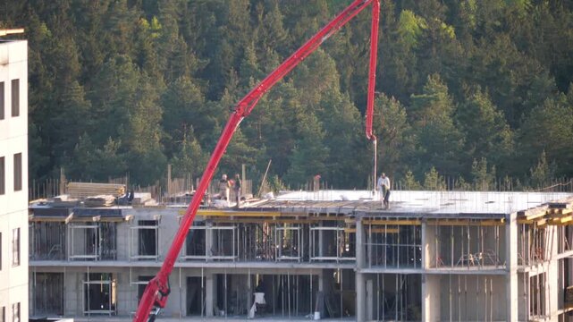 Construction workers pouring wet concrete mix on the top floor of a new residential building using a long concrete pump arm. Builder levels concrete to solidify into a reinforced concrete structure