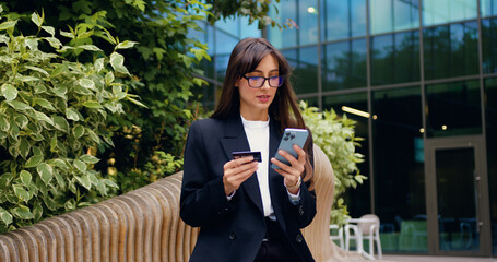 Young woman using phone and credit card for online banking while sitting near a glass office...