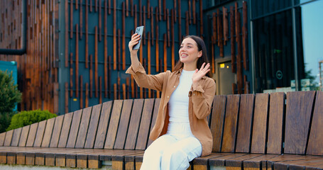 Close up of smiling Caucasian businesswoman talking on video call using phone outdoors while sitting on bench near office building. Online chat