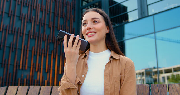 Young woman holding in hand smart phone talking with friend distantly uses easy voice messaging, concept of modern Ai technology, voice recognition, online translator over modern building background