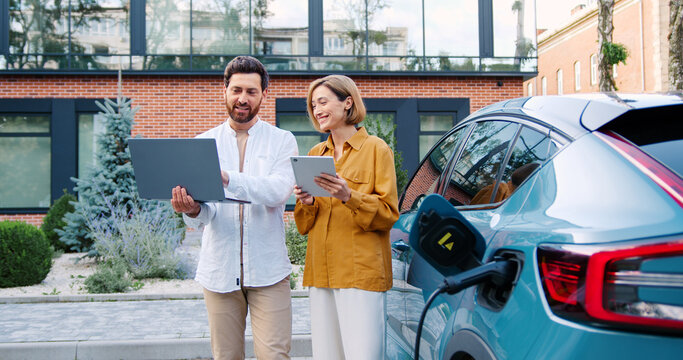 Male and female business team in elegant suits with laptop and tablet near electric car parking, modern office building in background