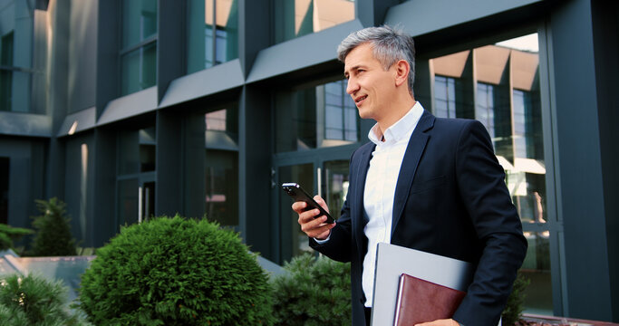 Man going outdoor with mobile phone in his hands. Middle aged businessman walking using smartphone outdoors in city near office building.