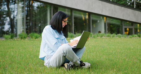 Focused young woman sitting alone on green grass in a sunny campus park, studying with a laptop and writing in a notebook. Freelance professional or university student working on a project outdoors