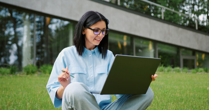 Young Caucasian woman in casual clothes sitting cross-legged on green grass in an urban park, concentrating on her laptop and balancing work with nature