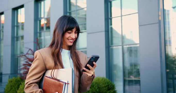 Young Caucasian woman confidently walking near a modern glass office building, smiling as she carries a laptop and a notebook in her hands, while checking her smartphone.