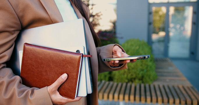 Close-up of woman's hands holding laptop and notebook in one hand while typing message on her smartphone with the other hand, outside