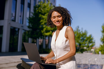 woman confidently working on laptop amid sunny outdoor environment with urban architecture