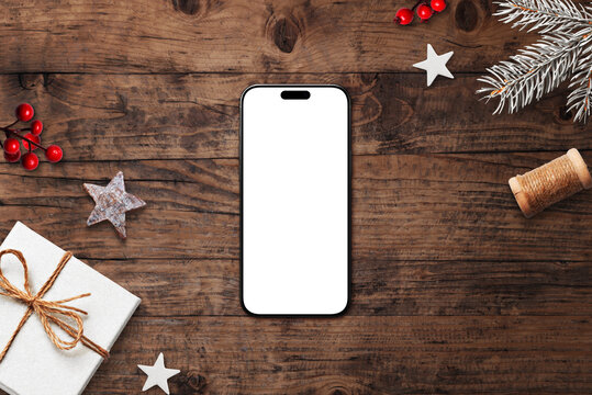 Smartphone with blank white screen on rustic wooden background, surrounded by Christmas decorations, pine branches, and festive ornaments