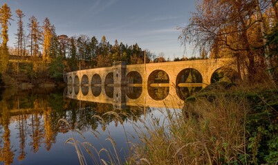 View of the Sedlice Dam (Vysočina, CZ) from the surface, reflection of the stone dam in the water, soft light of the setting November sun, yellow leaves on the trees