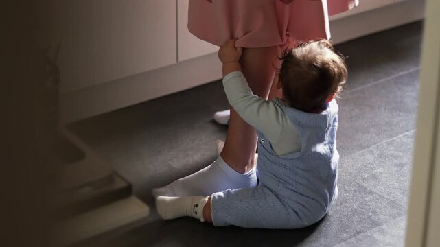 Baby sitting on floor holding mother&rsquo;s legs in kitchen, expressing attachment and need for closeness, family life and early emotional bonding