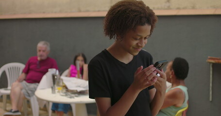 Teenager of African descent holding smartphone with green screen, chroma key feature displayed, smiling outdoors at a casual gathering with diverse adults in the background