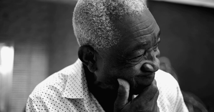 Elderly Afro-descendant man in black and white portrait, eyes closed with a pensive and slightly concerned expression, hand resting on chin in a reflective home setting