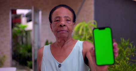 Elderly African American woman holding smartphone forward, green screen Chroma Key on display, blurred face in background, concept of digital technology and communication outdoors