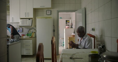 One Elderly Black man sitting at dining table in a cozy kitchen, concentrating on a smartphone, capturing the essence of daily routine and technology in a relaxed home environment