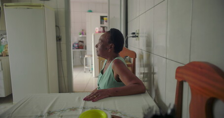 Elderly woman sitting alone at a dining table in a simple kitchen, staring thoughtfully into the distance, surrounded by appliances and tiled walls, reflecting a quiet moment