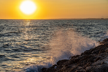 Obraz premium A minimalist sunset seascape where a wave crashes against rocky shore, sparkling with backlight as the sun hangs low over the shimmering silver Mediterranean Sea. Side, Antalya, Turkey.