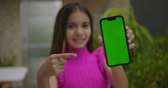 Young girl smiling and pointing at smartphone with green screen and chroma key in an outdoor setting, emphasizing customizable display for digital content or advertising