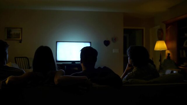 Group of people watching television together on a sofa in a dim living room at night, faces silhouetted by the screen glow, cozy movie night and streaming entertainment, family or friends bonding