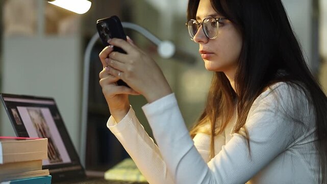 Woman seated at a library desk references a smartphone near a laptop, hybrid research workflow, productivity and end-of-day study atmosphere.