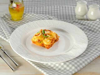 A portion of fresh lasagna rests on a textured white plate. Silver cutlery and condiments sit alongside, ready for the start of a delicious meal on a gingham tablecloth