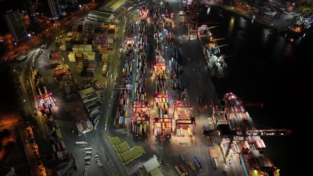 Bird's eye night view of a bustling industrial harbor in mersin, turkey, with illuminated gantry cranes loading and unloading colorful shipping containers from docked cargo vessels