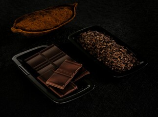 Dark chocolate bars rest on a tray, next to a pile of cocoa nibs and a split-open cocoa pod, all arranged on a textured dark surface. A spotlight emphasizes the rich colors