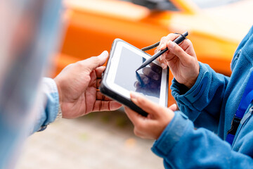 Person using a tablet to record information while another person hands over an item in an outdoor setting with an orange vehicle in the background