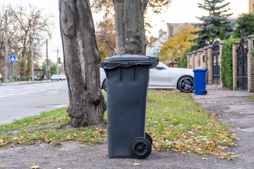 Obraz premium Black trash bin on the street beside a tree during autumn with fallen leaves and parked cars in the background