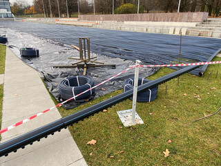 Construction site with black PVC pipes for refrigerant laid on vapor barrier. Pipes are connected to main header, forming cooling loop for outdoor seasonal ice rink.