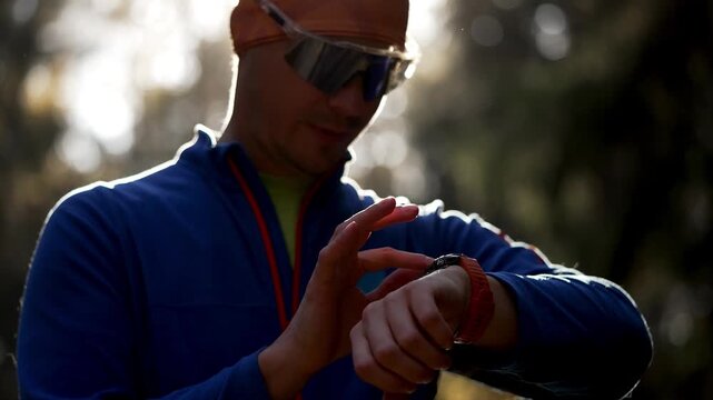 Athletic man in sportswear using a smart watch to check his workout data after running in a park, with the sun creating a beautiful lens flare and rim light around his silhouette - Powered by Adobe