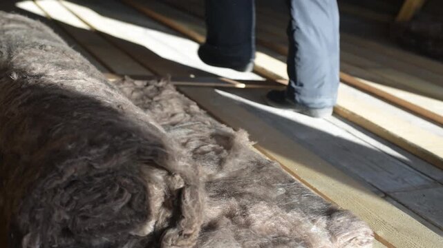 close-up shot of hands wearing gloves working with rolls of insulation material, in a construction or renovation setting. insulation appears fibrous and is being cut or manipulated.