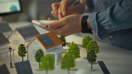 A design engineer is using a calculator and pen to assess data while planning solar panel installations on model houses. This activity takes place in a workspace focused on renewable energy solutions - Powered by Adobe