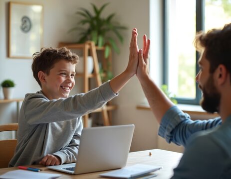 Father and son doing homework together, high five celebrates success. Boy smiles, dad helps with laptop learning, family bonding, school achievement.