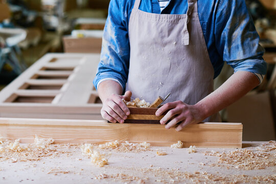 Caucasian young adult man working with wood planer, shaping wooden plank on workbench, hands covered in sawdust, focused on carpentry task in woodworking workshop environment