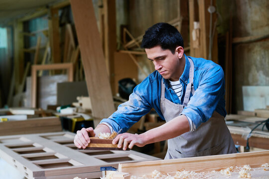 Caucasian young adult man working in carpentry workshop using hand planer on wooden board, focused on shaping wood, surrounded by woodworking tools and unfinished furniture pieces