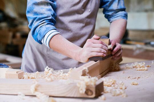 Caucasian young adult man using hand planer shaping wooden board in carpentry workshop, hands covered in sawdust, focusing on woodworking process, wood shavings scattered on table