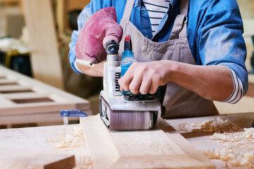 Caucasian young adult man using electric planer shaping wooden board in workshop, wearing apron and gloves, focusing on precise woodworking, wood shavings scattered on table