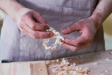 Caucasian young adult man shaping wood shavings with hands, standing at workbench, showing close up of skilled craftsmanship and detailed manual woodworking process