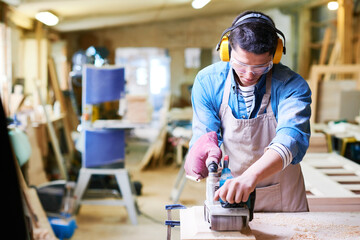 Young adult Caucasian man operating electric planer on wooden board in carpentry workshop, wearing protective goggles and earmuffs, focusing on woodworking project, hands guiding tool precisely